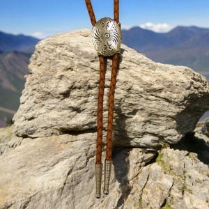 May include: A brown leather bolo tie with a silver-toned oval pendant featuring a decorative floral design. The tie is displayed against a rocky background.