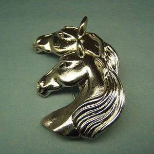 May include: Silver-toned brooch featuring two horses' heads facing opposite directions. The horses' manes are detailed and flowing.
