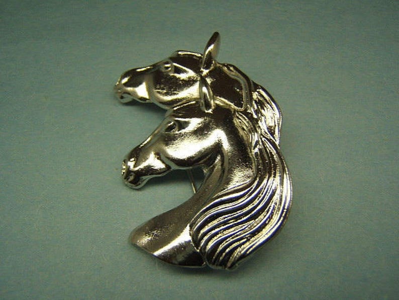 May include: Silver-toned brooch featuring two horses' heads facing opposite directions. The horses' manes are detailed and flowing.