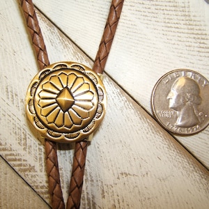May include: A brown leather bolo tie with a gold-toned metal bolo slide featuring a geometric design. The slide has a raised, diamond-shaped center surrounded by radiating lines.
