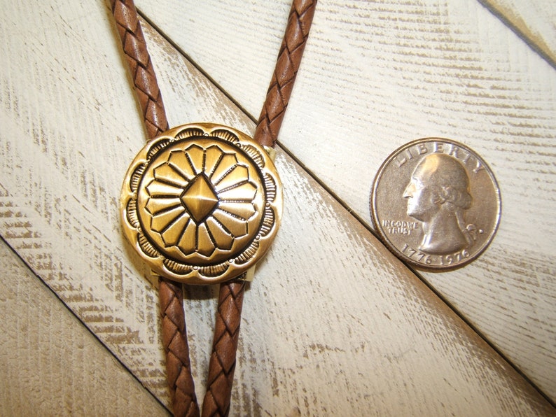 May include: A brown leather bolo tie with a gold-toned metal bolo slide featuring a geometric design. The slide has a raised, diamond-shaped center surrounded by radiating lines.