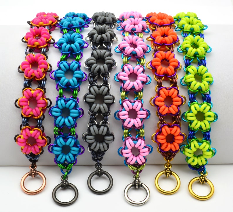 May include: Seven colourful bracelets with flower-shaped links. The bracelets are made of metal and have a variety of colours, including pink, blue, grey, orange, and green. Each bracelet has a small ring at the end.