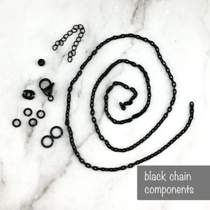 Pode incluir: Uma variedade de componentes de corrente pretos, incluindo uma corrente longa, uma corrente mais curta, argolas, um fecho e outros pequenos conectores. Os componentes est&atilde;o dispostos sobre uma superf&iacute;cie de m&aacute;rmore branco e cinza. O texto "black chain components" est&aacute; numa caixa cinza.