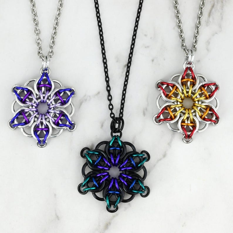Pode incluir: Tr&ecirc;s colares com pingentes de flores em malha de corrente. Um &eacute; prateado com detalhes roxos e azuis, outro &eacute; preto com verde-azulado e roxo, e o &uacute;ltimo &eacute; prateado com detalhes vermelhos, laranjas e amarelos.