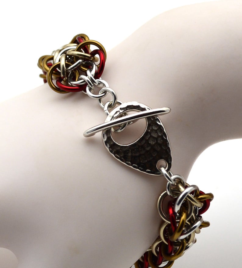 Puede incluir: Un brazalete de malla de cadena roja, dorada y plateada con un cierre de palanca de plata martillado.