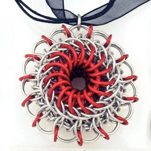 May include: A silver and red chainmail pendant in the shape of a flower. The pendant is made of interlocking metal rings and has a hole in the center.
