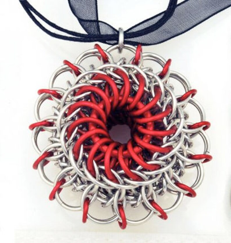 May include: A silver and red chainmail pendant in the shape of a flower. The pendant is made of interlocking metal rings and has a hole in the center.