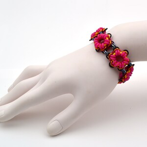 May include: A pink and yellow rubber band bracelet with a flower design. The bracelet is on a white mannequin hand.