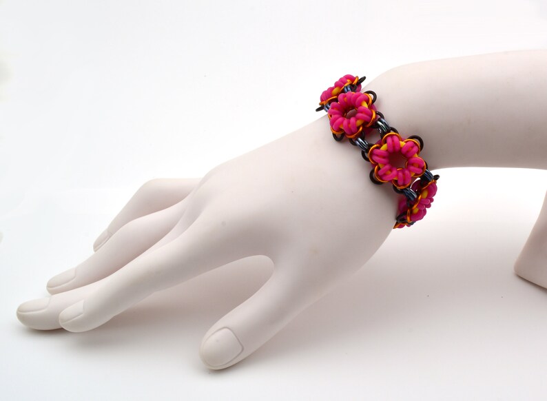 May include: A pink and yellow rubber band bracelet with a flower design. The bracelet is on a white mannequin hand.