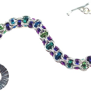 Puede incluir: Un brazalete plateado con una cadena colorida hecha de peque&ntilde;os anillos de metal. La cadena es una mezcla de morado, verde y azul. El brazalete tiene un cierre plateado y un anillo plateado con una superficie texturizada.