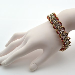 Puede incluir: Un brazalete de malla de cadena plateado y dorado con detalles rojos. El brazalete est&aacute; hecho de peque&ntilde;os anillos de metal que se entrelazan para crear un patr&oacute;n de malla de cadena.