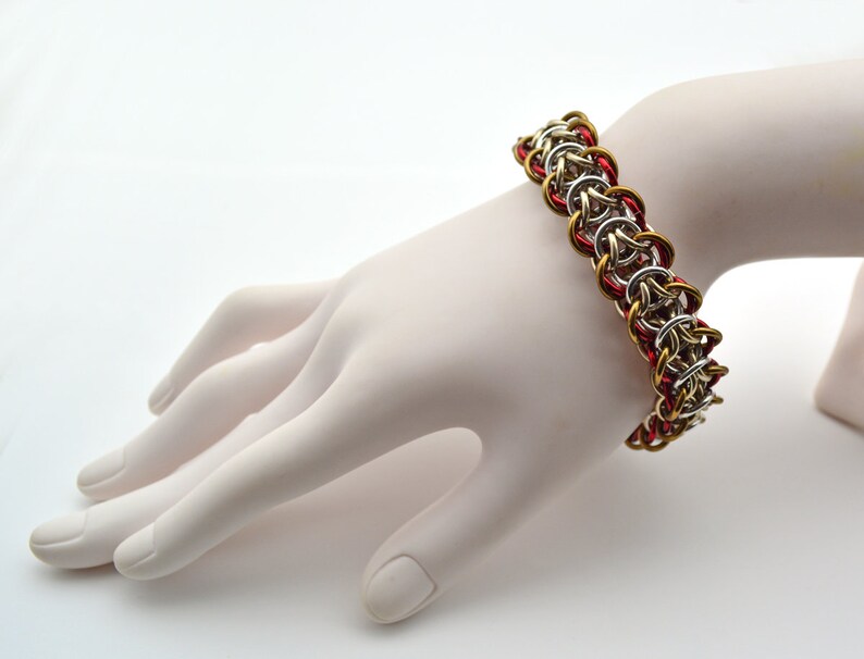 Puede incluir: Un brazalete de malla de cadena plateado y dorado con detalles rojos. El brazalete est&aacute; hecho de peque&ntilde;os anillos de metal que se entrelazan para crear un patr&oacute;n de malla de cadena.