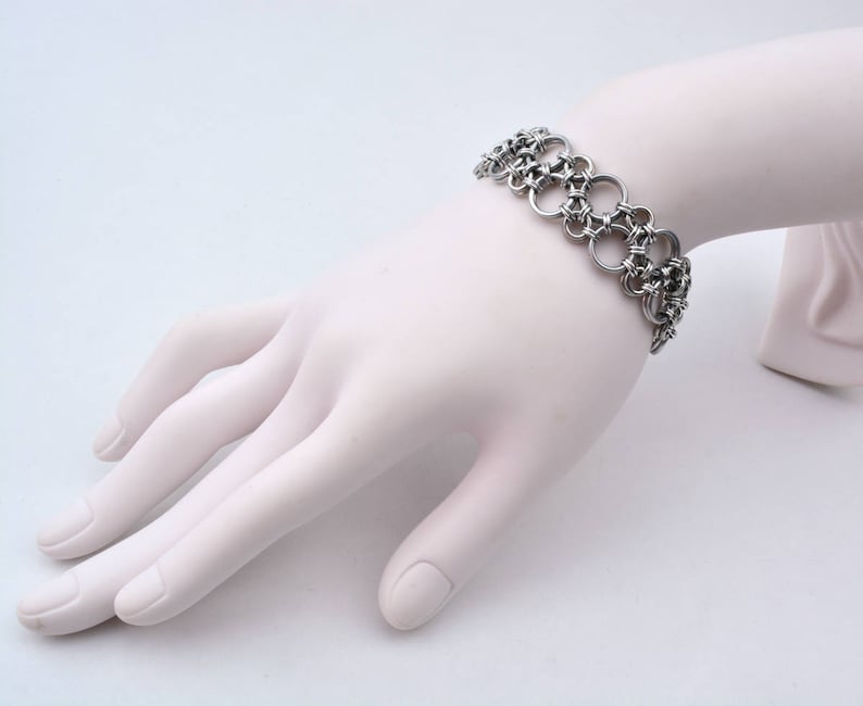 May include: A silver chainmail bracelet with a woven design. The bracelet is on a white mannequin hand.