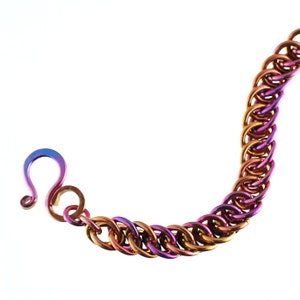 May include: A chain bracelet made of titanium with a gradient of purple, pink, and gold colors. The bracelet has a hook clasp.