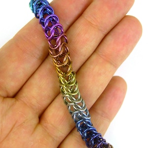 May include: A rainbow colored metal chainmail bracelet. The bracelet is made of small, interlocking metal rings that are woven together in a pattern. The bracelet is a gradient of colors, starting with blue at one end and transitioning to purple, then gold, and finally silver at the other end.