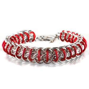 May include: A silver and red chainmail bracelet with a clasp. The bracelet is made of small, interlocking metal rings.
