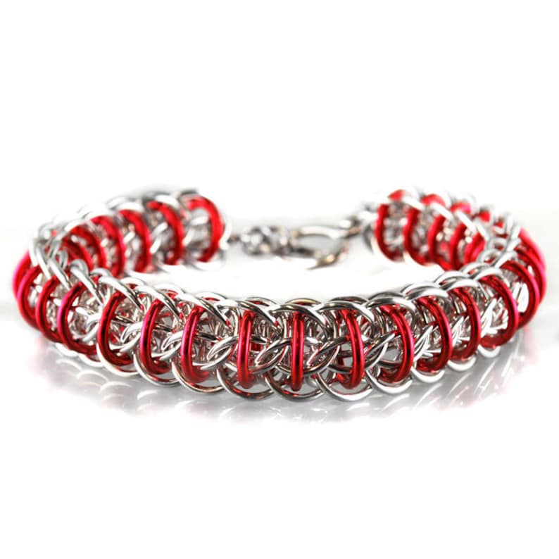 May include: A silver and red chainmail bracelet with a clasp. The bracelet is made of small, interlocking metal rings.