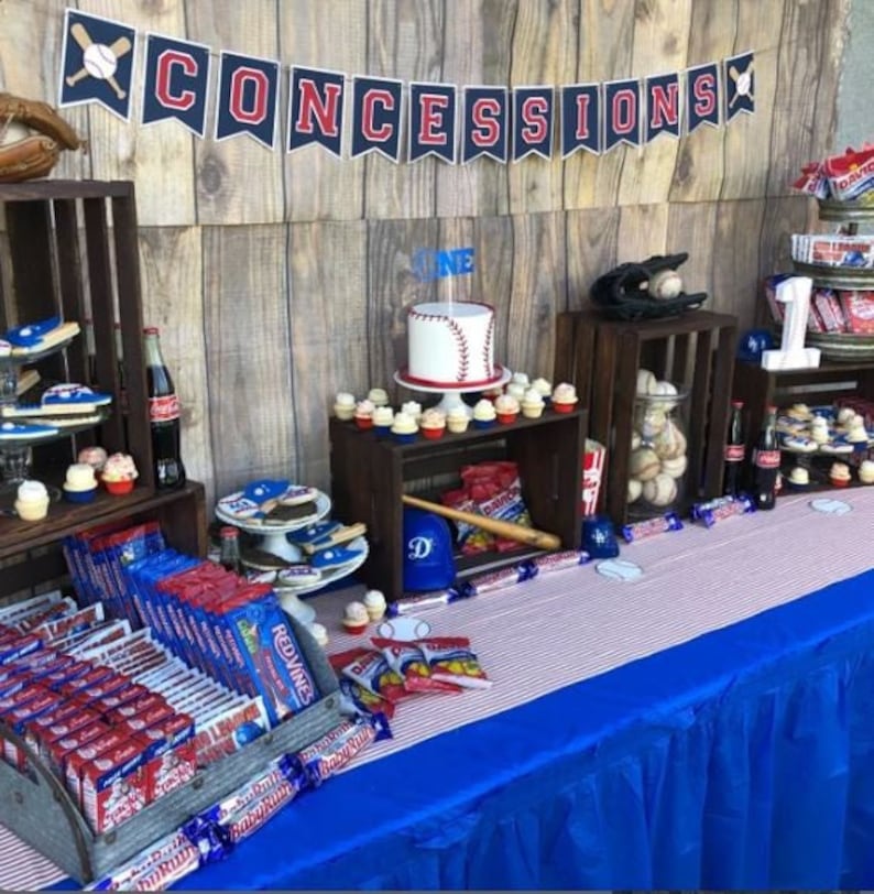 May include: A baseball-themed party table setting with a blue and white striped tablecloth, a blue tablecloth, and a wooden crate display. The table is decorated with baseball-themed treats, including cupcakes, cookies, and candy. The table also features a baseball-themed cake with a baseball on top and the number "one" on the side. The table is decorated with a banner that says "CONCESSIONS".