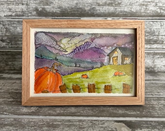 Puede incluir: Una pintura en acuarela enmarcada que representa un paisaje con una calabaza naranja grande en primer plano, una cerca de madera, una casa pequeña y colinas bajo un cielo morado y gris con patrones de nubes en un marco de madera.