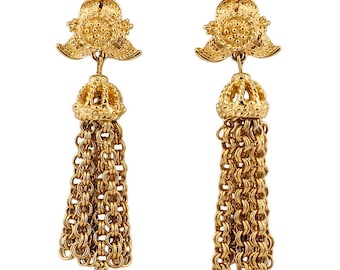May include: A pair of gold-tone dangle earrings featuring a textured floral top component connected to a domed cap. Below the cap, multiple strands of interlocking circular gold-tone metal rings form a tassel design that hangs vertically.