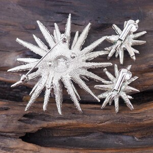 May include: A silver starburst brooch and matching earrings. The brooch has a pin back and the earrings have post backs. The jewelry is made of metal and has a sparkly finish.