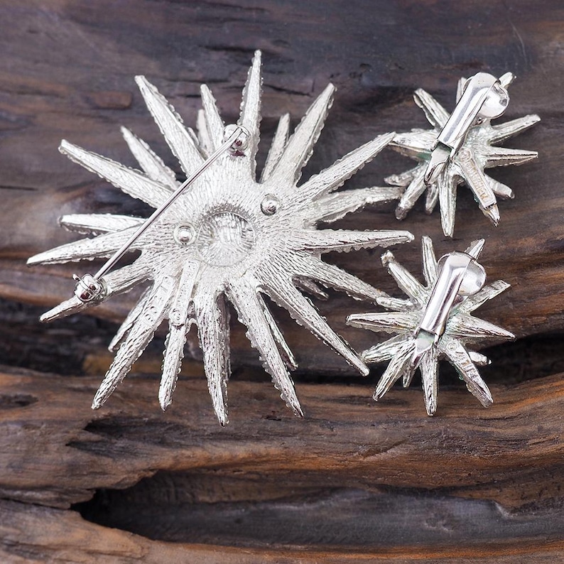 May include: A silver starburst brooch and matching earrings. The brooch has a pin back and the earrings have post backs. The jewelry is made of metal and has a sparkly finish.