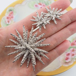 May include: A pair of silver tone brooch pins in the shape of a starburst. The pins are made of textured metal and have a three-dimensional design.