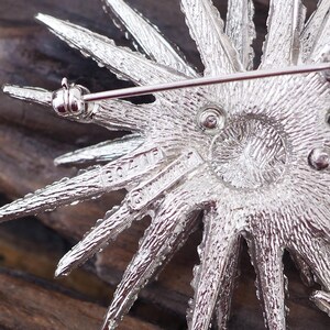 May include: Silver tone brooch with a starburst design. The back of the brooch has the words "Bozell" and "Guaranteed" inscribed on it.