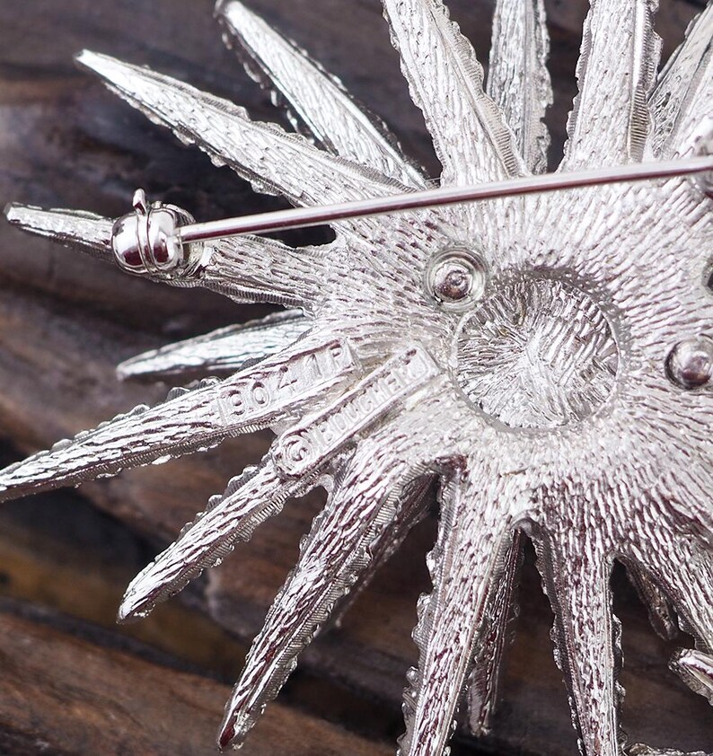 May include: Silver tone brooch with a starburst design. The back of the brooch has the words "Bozell" and "Guaranteed" inscribed on it.