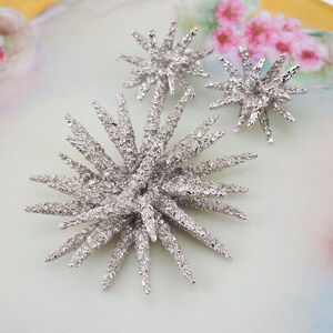 May include: A set of three silver starburst brooches with a textured finish. The brooches are arranged on a white background.