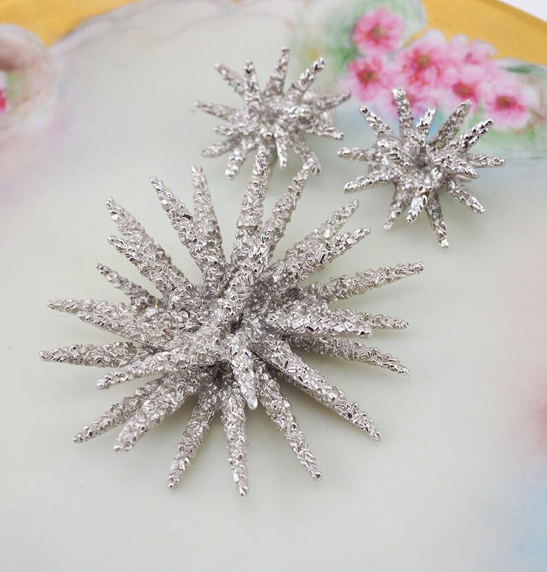 May include: A set of three silver starburst brooches with a textured finish. The brooches are arranged on a white background.