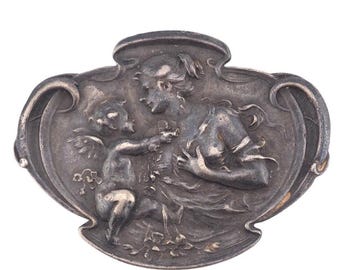 May include: Antique silver-toned brooch featuring a bas-relief design. The brooch depicts a woman and a cherub, surrounded by floral elements, within an ornate, oval frame. The metal has a tarnished appearance.