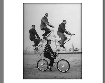May include: Black and white framed photograph of a group of people riding a multi-tiered bicycle. Four people are seated on the upper levels, while one person pedals the base bicycle. The photograph is set against a neutral background.