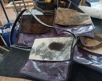 May include: A stack of four brown and white cowhide leather purses with clear plastic panels. The purses have a brown leather strap.