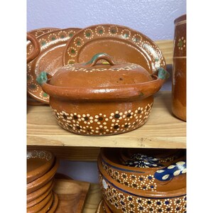 May include: A terracotta casserole dish with a lid, featuring a white and brown dotted pattern. The dish has a green handle and is displayed on a wooden shelf with other terracotta dishes.
