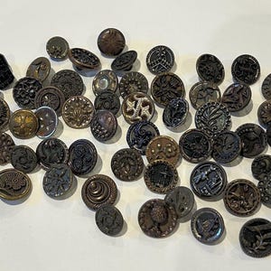Puede incluir: Una colección de botones antiguos de varios tamaños y diseños. Los botones son principalmente de color marrón oscuro y negro, con intrincados patrones en relieve, incluidos motivos florales, geométricos y figurativos. Algunos botones tienen un brillo metálico.