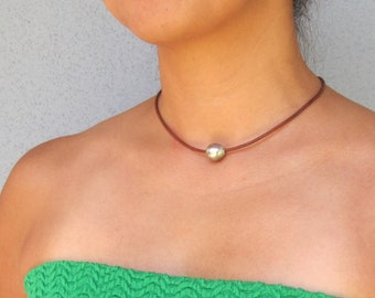 May include: A simple necklace featuring a brown leather cord and a large, silver-colored sphere pendant. The necklace is worn against a green strapless top. The background is a plain, light gray wall.