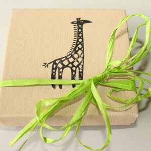 K&ouml;nnte beinhalten: Eine quadratische, beige Geschenkbox mit einer schwarzen Giraffenillustration auf der Oberseite. Die Box ist mit einem hellgr&uuml;nen Bastband gebunden. Das Giraffen-Design ist ein einfacher, stilisierter Druck.