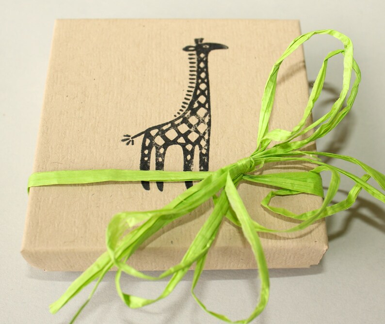 K&ouml;nnte beinhalten: Eine quadratische, beige Geschenkbox mit einer schwarzen Giraffenillustration auf der Oberseite. Die Box ist mit einem hellgr&uuml;nen Bastband gebunden. Das Giraffen-Design ist ein einfacher, stilisierter Druck.
