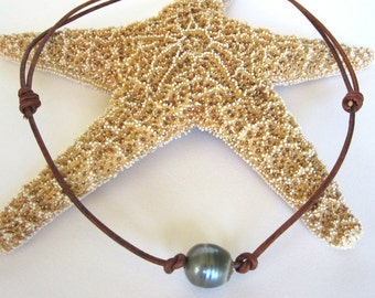 May include: A dark gray pearl necklace with a brown leather cord. The cord is knotted on each side of the pearl and at the ends for adjustment. The necklace is displayed on a starfish.
