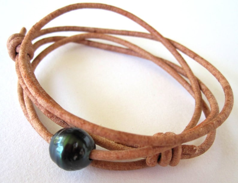 K&ouml;nnte beinhalten: Ein Wickelarmband mit einer dunklen, gl&auml;nzenden schwarzen Perle, die auf einer hellbraunen Lederschnur aufgef&auml;delt ist. Die Schnur ist an mehreren Stellen geknotet, wodurch ein geschichteter Effekt entsteht. Das Armband ist zum Wickeln um das Handgelenk konzipiert.