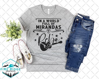 May include: A heather gray t-shirt with the text "IN A WORLD FULL OF MIRANDAS BE A Reba" in black, with a guitar graphic. The shirt is paired with blue jeans and white sneakers. A small plant and a logo are also visible.