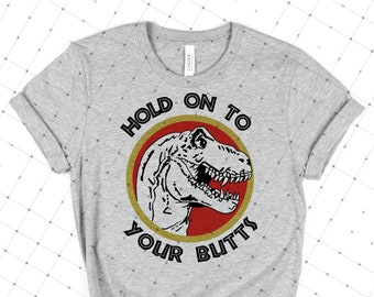 Puede incluir: Camiseta gris claro con una ilustración de un Tyrannosaurus Rex dentro de un círculo rojo y dorado. El texto "HOLD ON TO YOUR BUTTS" está impreso encima y debajo de la imagen del dinosaurio en negrita negra.