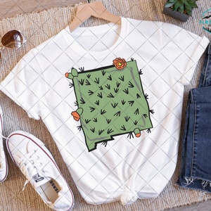 Peut inclure: T-shirt blanc avec un motif de cactus vert, des fleurs oranges et des épines. Le t-shirt est associé à un jean bleu, des baskets blanches et des lunettes de soleil. Le motif est une illustration graphique.