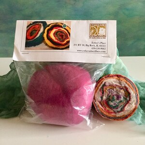 May include: A close-up of a package containing two felted wool balls. One ball is a solid pink color, and the other is a multi-colored swirl of wool. The package is labeled with the name "Esther's Place" and a website address.