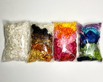 May include: Six bags of colorful curly wool roving. The bags are arranged in a row from left to right: white, brown, orange, pink, blue, and green.
