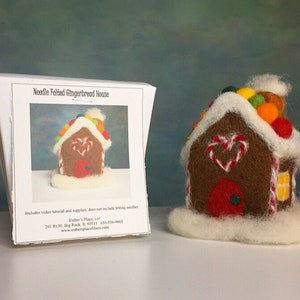 May include: Needle felted gingerbread house kit. The kit includes a video tutorial and supplies. The gingerbread house is brown with white icing and red candy hearts. The roof is white with colorful candy balls. The house is sitting on a white base.