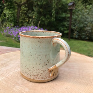 May include: A handmade ceramic mug with a pale green glaze and speckled brown details. The mug has a unique handle design and a brown rim. The mug is sitting on a wooden surface.