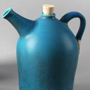 May include: A teal ceramic jug with a spout and handle, topped with a cork stopper. The jug has a speckled texture and a light tan base. The handle has a gold accent.