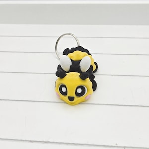 May include: A yellow and black bee-shaped keyring with a silver ring. The bee has white antennae, pink cheeks, and large black eyes. A charming, whimsical accessory.